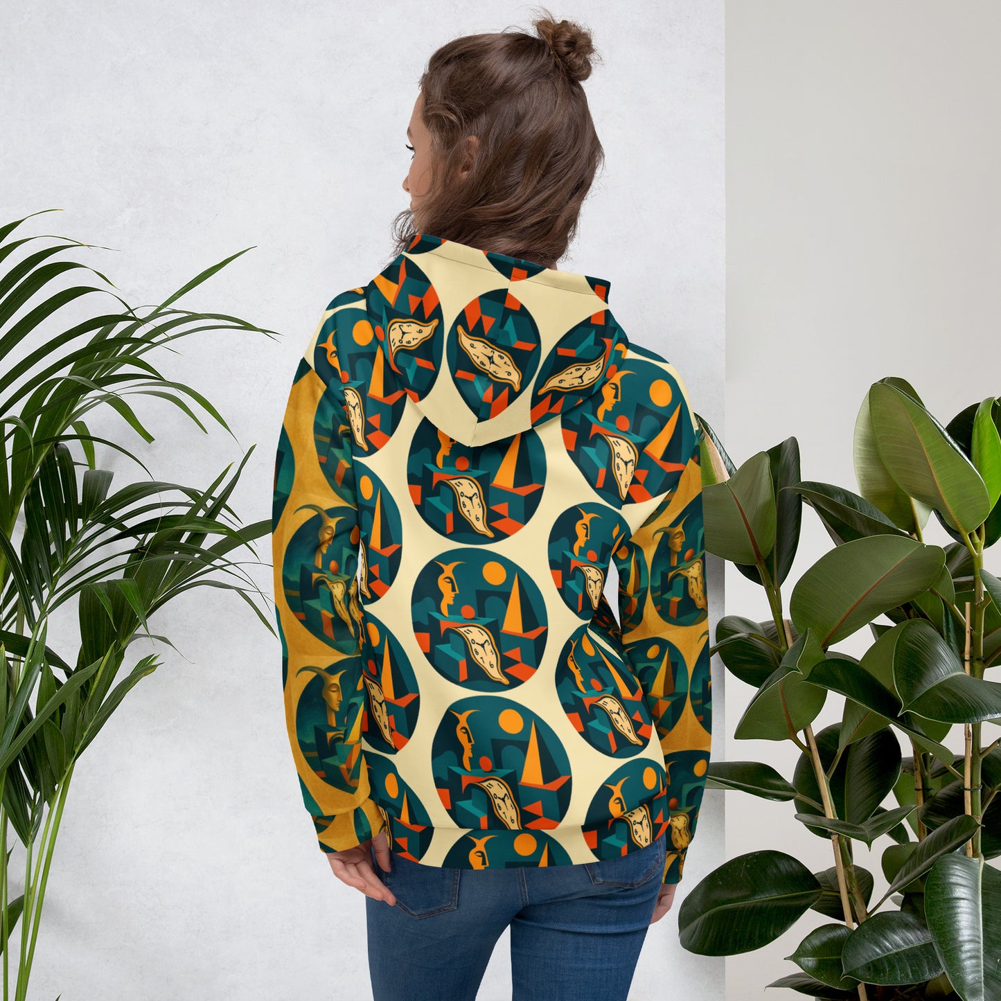 Person wearing a colorful patterned hoodie standing between two plants against a white wall.