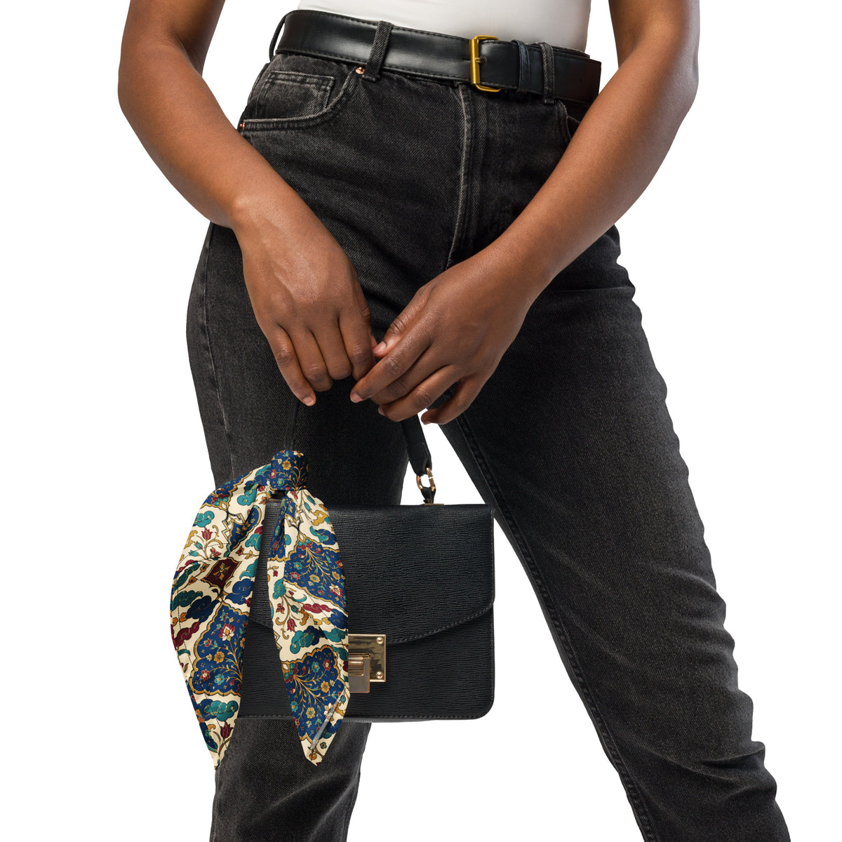 A person wearing black jeans and a black belt holds a black handbag with a colorful patterned scarf tied to the handle, against a plain white background.