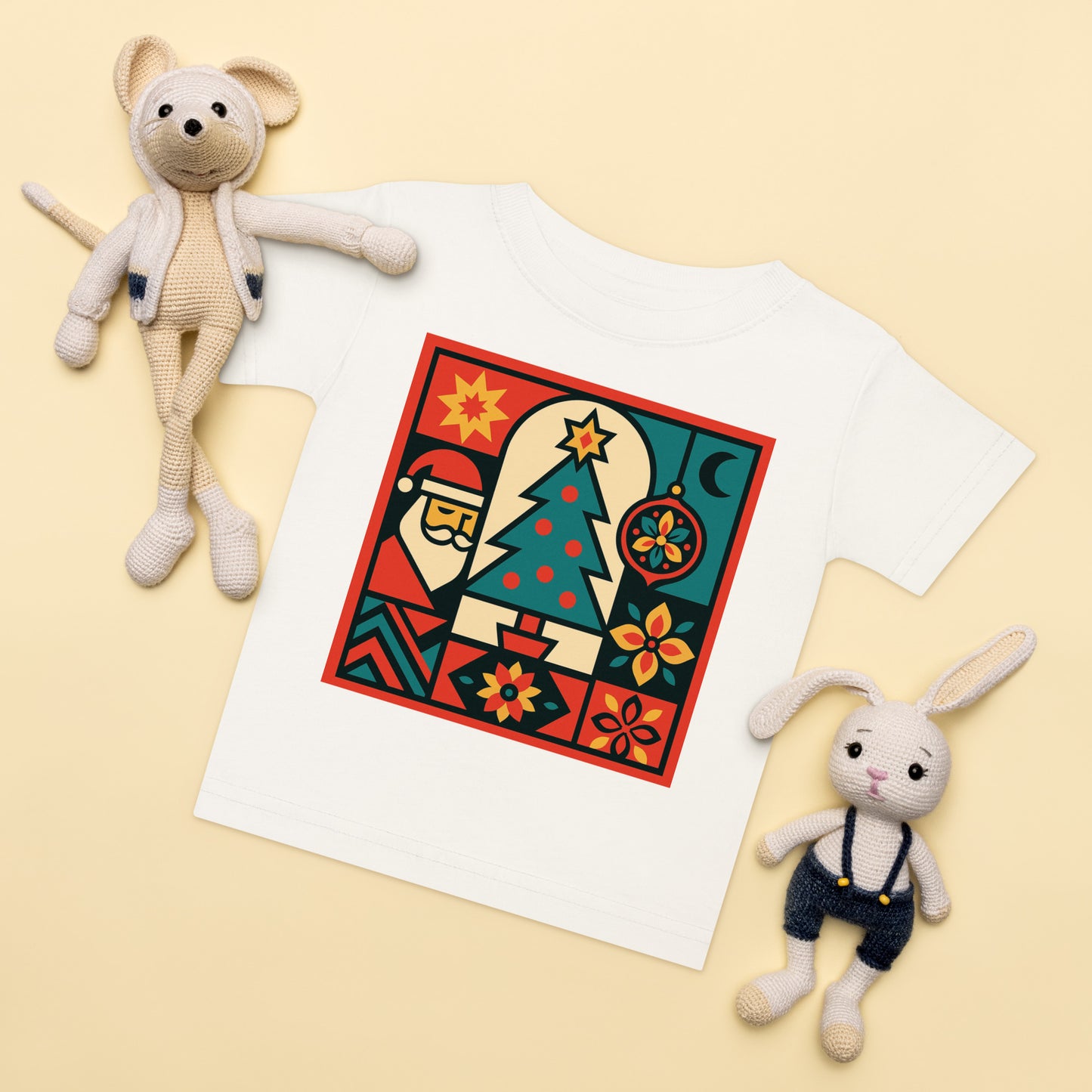 White t-shirt with a colorful graphic design featuring a Santa Claus-like figure, tree, and flowers, placed on a beige surface with two stuffed toys.