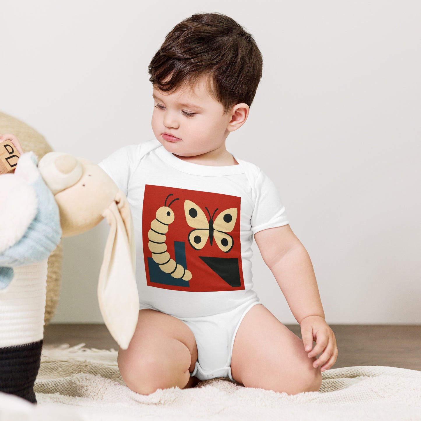 Child wearing a onesie with a butterfly and caterpillar design, sitting on a carpeted floor.